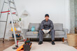© pichet - Young technician in work uniform sitting on couch at home with tools, ladder, and equipment, preparing for repair or maintenance tasks indoors