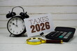 © may1985 - Taxes 2026 text on paper card with alarm clock, calculator and magnifying glass on wooden background