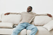 © SHOTPRIME STUDIO - Relaxed young Black man enjoying leisure time on a beige couch, wearing a cozy sweater and jeans, exudes happiness in a modern living room setting