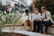 © qunica.com - Three colleagues collaborate on a project outdoors, while two others discuss nearby. The scene portrays teamwork and professional interaction in a modern city setting.