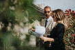 © qunica.com - Two colleagues, one in formal attire and the other in sunglasses, having a professional discussion with documents in an outdoor environment surrounded by green foliage.