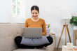 © NAMPIX - A happy young woman sits comfortably on a sofa, smiling as she works on a laptop. The cozy living room setting suggests she is working from home,