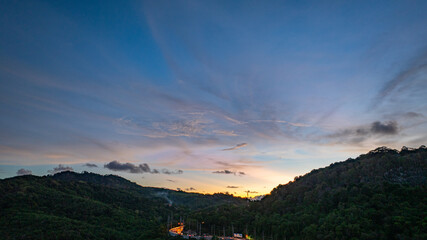 Wall Mural - Aerial hyper lapse view of A breathtaking sunset paints the sky in vivid hues of orange and blue over a coastal town, with lush green hills framing the scene. cloud in dramatic sunset over the ocean