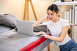 © bongkarn - Asian woman hand supporting head looking at laptop while sitting leaning against sofa bed on floor.