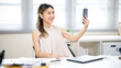 © bongkarn - Pretty woman office worker holding smartphone taking a selfie or video calling at table in office.
