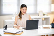 © bongkarn - Asian woman office worker holding pen and looking at laptop while sitting at white table in office.