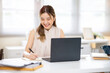 © bongkarn - Happy asian woman office worker holding pen writing in notebook with laptop on white table in office