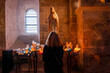 © sirbouman - Silent devotion of a woman with her back turned praying before the image of the Virgin Mary with votive candles inside a medieval stone church.