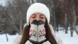 © ekaterinabyuksel - A portrait of a woman wearing a warm white knitted hat outdoors in winter. The young woman enjoys the beautiful weather and the first snow. Active winter recreation. She warms her hands with her