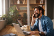 © Stockphotodirectors - A man with a beard sits at a wooden desk in his home office, engaged in a phone conversation while typing on his laptop. Bright light streams through the window, illuminating the room.