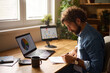 © Stockphotodirectors - A focused individual actively engages with a laptop and a tablet while writing notes in a cozy home office. A warm atmosphere is enhanced by sunlight streaming through the window.