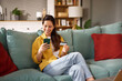 © Stockphotodirectors - A woman smiles while using her smartphone to make an online purchase. She holds a credit card in one hand, relaxed on a couch with colorful cushions in a sunlit living room.