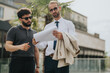 © qunica.com - Two well-dressed individuals engage in a discussion outdoors while examining paperwork in a modern urban environment, capturing a professional and collaborative work scenario in natural light.