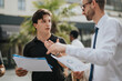 © qunica.com - Two business professionals engage in a discussion, carefully reviewing documents, outdoors near modern buildings.