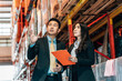 © BigBlues - A business man in a suit gestures toward the upper shelves in a warehouse as he talks to a business woman, who holds a clipboard and looks up.