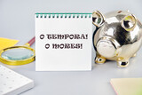 o tempora, o mores (O, the times O, the morals) Latin phrase on a white notebook on a gray background near a piggy bank, a calculator and stickers