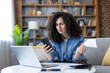 © Liubomir - Woman with curly hair checking her smartphone and documents, experiencing stress and confusion while managing personal finances and experiencing budgeting difficulties at home