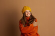 © Alliance - A cheerful woman in a yellow knit beanie and an orange turtleneck sweater, with arms crossed, smiles upwards. The warm tones and cozy attire evoke an inviting autumn or winter atmosphere.