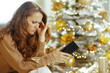 © Alliance - A woman with a distressed expression stares at her smartphone, resting her head on her hand, against a blurred, softly lit Christmas tree background, conveying a somber holiday mood.