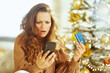 © Alliance - A middle-aged woman looks shocked at smartphone while holding credit card, facing unexpected expense or holiday spending concern. A blurred Christmas tree with warm lights sets anxious festive mood.