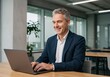 © shahida  - Smiling middle aged businessman in a dark suit and white shirt actively working on a laptop in a modern office setting