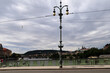 © LIS - Puente urbano con farola ornamental de doble esfera en primer plano, un río fluyendo entre edificios y zonas verdes, y colinas con una torre al fondo bajo un cielo gris.