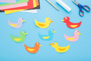 Wall Mural - Created and glued colorful duck shapes. Scissors, glue stick, pencil and application paper on light blue table background. Pastel color. Closeup. Child handcraft. Top down view.