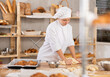 © JackF - Young female apprentice baker in white chef coat and hat kneading dough on floured table in artisan bakery, surrounded by freshly baked bread and pastries displayed on wooden shelves