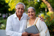 © Azmanrazak - Indian Elderly Couple Smiling Together in the Park, Holding a Tablet on a Sunny Day, Surrounded by Greenery and Flowers, Celebrating Happy Retirement