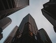 © QITIAN - Looking up at modern skyscrapers in an urban setting with a blue-gray sky.