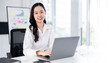 © NAMPIX - A happy young Asian woman sits at her desk, smiling warmly at the camera while working on a laptop.