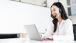 © NAMPIX - A happy, smiling young Asian woman wearing a headset, working on a laptop in a modern office or call center.