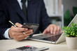 © khunkornStudio - Manager checks phone while typing on laptop in office for finance report email communication mobile banking productivity technology workflow remote work concept modern business.