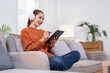 © NAMPIX - A happy young Asian woman, wearing wireless headphones, sits comfortably on a sofa and uses a stylus to work on a tablet.