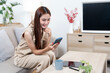 © NAMPIX - A happy young Asian woman in casual attire smiles while using her smartphone and a tablet. She is seated comfortably on a sofa in a modern and inviting living room.