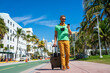 A tourist with camera and luggage on streets of Miami. A man tourist enjoys his summer vacation in a tropical city. A traveler tourist with suitcase on summer city. Man tourist walk on city.