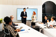 © artitwpd - Diverse team in a modern conference room during a presentation. Two colleagues stand by a large screen while others sit with laptops and notes in a corporate meeting.