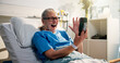 © peopleimages.com - Senior woman, phone and video call in hospital bed with wave, surprise or get well soon message. Person, smartphone and virtual contact on social media with rehabilitation, hello or chat at clinic