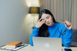 © Tj - Young Asian woman experiencing eye strain and headache while working from home on a laptop, feeling exhausted and stressed from prolonged screen time and overworking