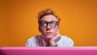 © Eve - Bored young man with red hair and glasses sitting at desk with pink laptop on orange background. Concept of procrastination, waiting, office routine, student life, boredom, stress and creativity.