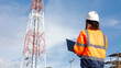 © Happy Photo - A worker in safety gear stands with a laptop, looking up at a tall telecommunications tower. The bright sky adds to the scene, showcasing a busy work environment