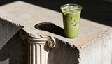 Iced matcha latte with ice in a clear plastic cup on a stone ledge with sunlight and shadow