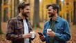 © miss irine - Two young bearded men talk, walk in autumn forest, hold coffee cups, using smartphone. Guys wear casual clothes, smiling, enjoy nature and friendship. Communication, autumn vibes, social media.