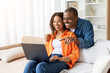 © Anastasiya - Smiling African American couple sitting on sofa with laptop, enjoying time together. Positive lifestyle, family bonding, love, technology and modern home atmosphere