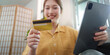 © Natee Meepian - Online Shopping: Young woman holding credit card and tablet, smiling at home