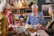 © AnnaStills - Group of senior Caucasian men and women sitting together enjoying tea and cake, engaging in conversation around table with desserts, smiling and interacting in cozy indoor setting