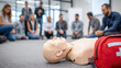 © dominic - First Aid training session with CPR dummy on floor surrounded by attentive participants engaged in emergency response education workshop indoors.
