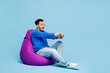 © ViDi Studio - Full body young man of African American ethnicity wear sweatshirt casual clothes sit in bag chair hold in hand play pc game with joystick console isolated on plain pastel light blue background studio.