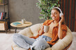 © ViDi Studio - Young fun woman wear orange casual clothes listen music in headphones drink coffee sits in chair stay at home hotel flat rest relax spend free spare time in living room indoor. People lounge concept.
