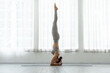 © 220 Selfmade studio - Asian Young woman practicing headstand yoga pose indoors with natural light, wearing gray sportswear, demonstrating balance and strength in calm, bright room